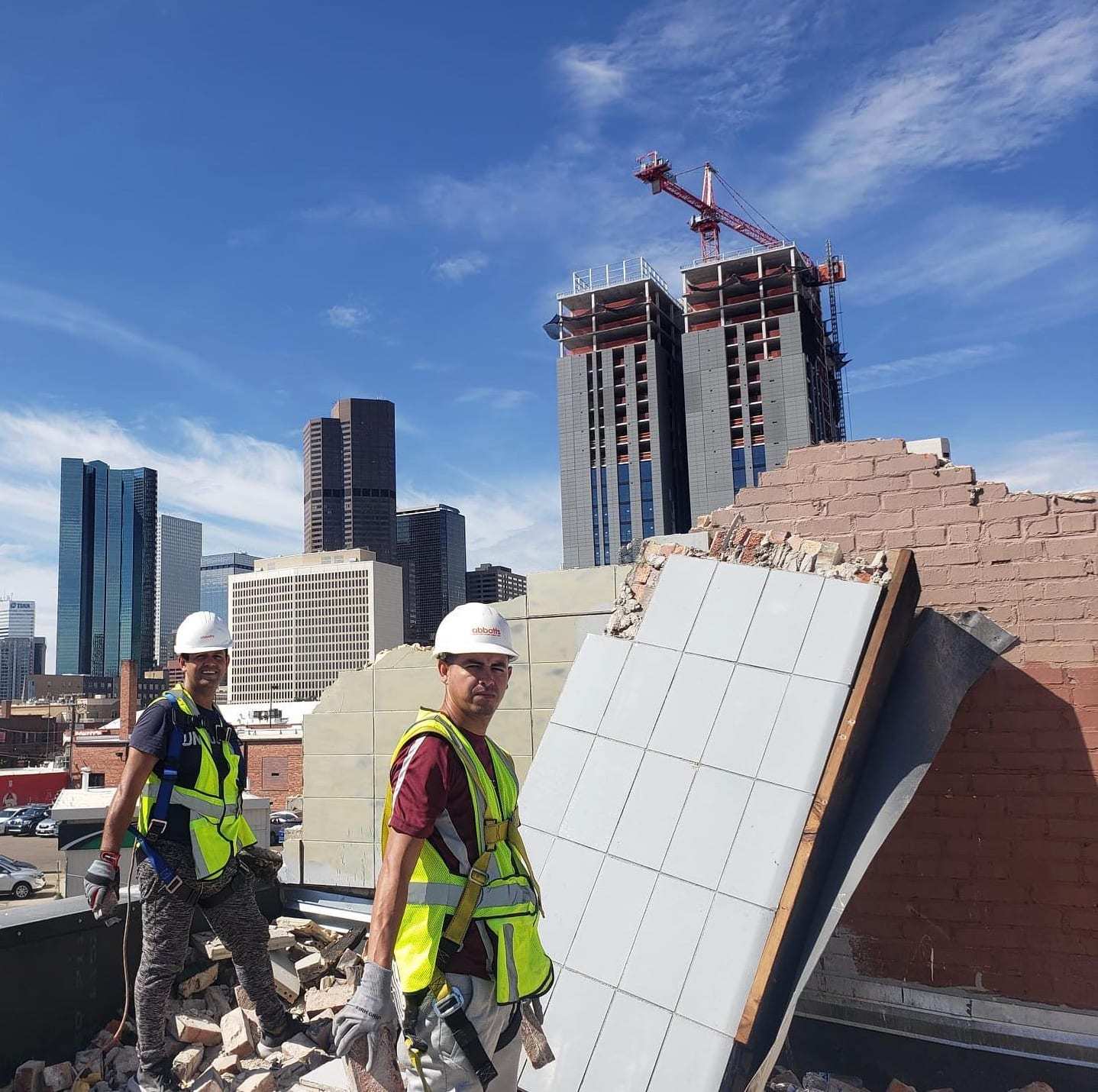 Two workers in hard hats on a demolished rooftop with the Denver skyline behind